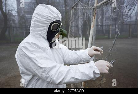 Vue latérale de l'écologiste en costume de rayonnement tenant les pinces et le tube à essai tout en prenant des échantillons dans la rue abandonnée, en portant un uniforme de protection, un masque à gaz et des gants. Concept de pollution de l'environnement. Banque D'Images