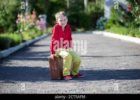 Un garçon triste est assis sur une vieille valise sur la route. L'enfant est sur le point de partir. Banque D'Images