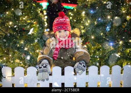 Bel enfant dans la rue de Noël. Un garçon en vêtements d'hiver sur fond d'arbres de Noël et de neige. Banque D'Images