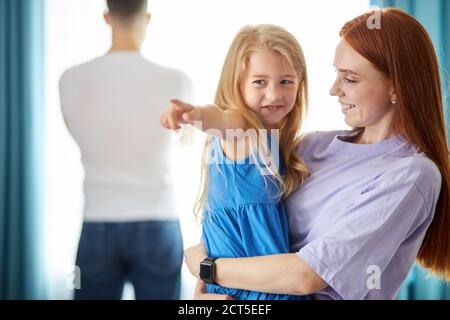 redhead caucasien fille tenir la fille dans les mains à la maison, ils sourire, mari offensé ne reçoit pas l'attention d'eux, tourné à la fenêtre Banque D'Images