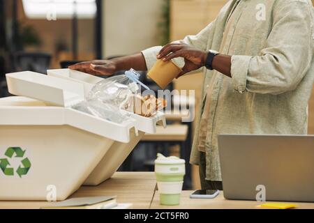 Portrait rogné d'un homme afro-américain méconnaissable mettant une tasse de papier dans le bac de tri des déchets dans le bureau, espace de copie Banque D'Images