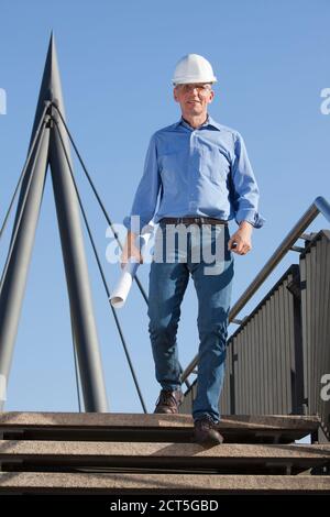 Architecte ou ingénieur souriant avec casque et plan sa main marchant au rez-de-chaussée dans un chantier de construction - foyer sur la personne Banque D'Images