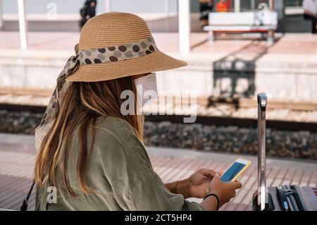 Vue latérale d'une femme méconnue sous un chapeau et un masque assis sur le banc près de la valise et de la navigation de smartphone sur la gare pendant une pandémie Banque D'Images