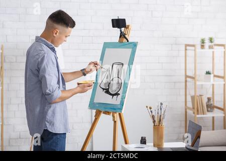 Passe-temps d'art et leçon en ligne pendant l'épidémie de COVID-19. Jeune homme dessine une image abstraite sur un chevalet avec un smartphone près de la table avec des brosses et un ordinateur portable Banque D'Images