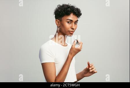 Jeune homme appliquant du brillant à lèvres. Homme liquide de genre mettant sur le maquillage sur ses lèvres avec applicateur contre fond blanc. Banque D'Images