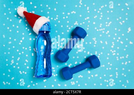 Appartement de sports de Noël avec haltères et bouteille d'eau dans un chapeau de père Noël rouge sur fond bleu. Concept de Noël et de vacances du nouvel an pour la remise en forme, W Banque D'Images