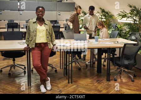 Portrait complet d'une jeune femme afro-américaine regardant un appareil photo debout, debout sur un bureau moderne, avec des personnes travaillant en arrière-plan, dans un espace de copie Banque D'Images