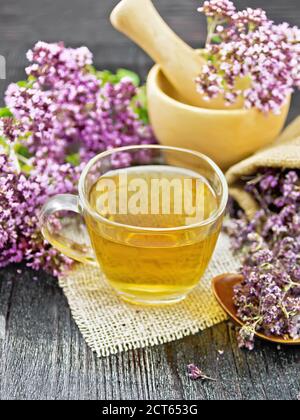 Tisane d'origan dans une tasse de verre sur toile de jute, fleurs fraîches dans le mortier et sur la table, fleurs de marjolaine séchées dans un sac et une cuillère sur planche en bois sombre Banque D'Images