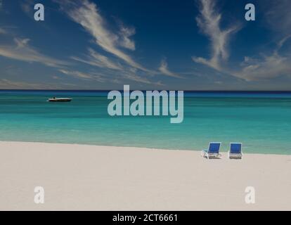 Une plage tropicale déserte avec deux chaises longues vides, la mer des Caraïbes en arrière-plan et un bateau de luxe amarré près de la plage de sable blanc. La perfection Banque D'Images