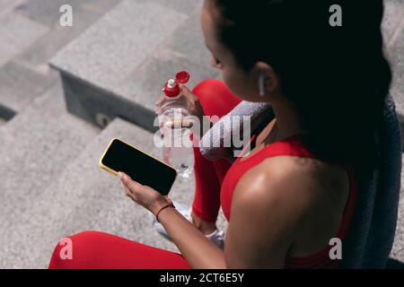 Les jeunes femmes ont des cheveux foncés dans des vêtements de sport rouges et les écouteurs blancs tiennent le smartphone et la bouteille d'eau. Vue de coupe à angle élevé. Serviette violette sur elle Banque D'Images