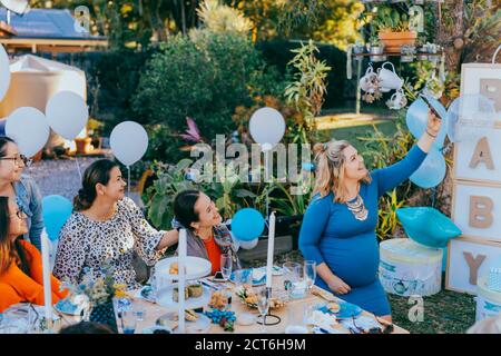 Des amies qui prennent le selfie avec une femme enceinte sous une douche de bébé. Photographie mobile, décorations de fête en blanc et bleu, bébé garçon Banque D'Images