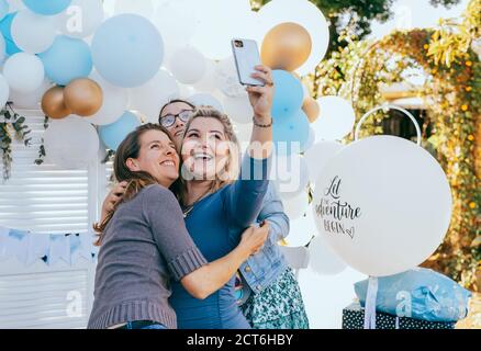 Des amies qui prennent le selfie avec une femme enceinte sous une douche de bébé. Photographie mobile, décorations de fête en blanc et bleu, bébé garçon Banque D'Images