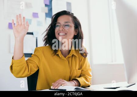 Une jeune employée de bureau gaie a soulevé la main pour saluer quelqu'un. Fille positive en vêtements jaunes avec verres ronds. Travail ou travail de bureau Banque D'Images