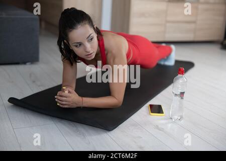 Jeune femme en survêtement rouge faisant de l'exercice ou du yoga à la maison. Sérieuse concentrée bien construite fille debout seul en position de planche sur le tapis de yoga Banque D'Images