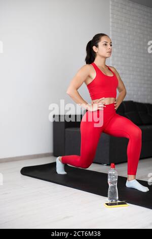 Jeune femme en survêtement rouge faisant de l'exercice ou du yoga à la maison. Vue latérale de la jeune fille debout sur une jambe position squat tenant les mains sur les hanches Banque D'Images