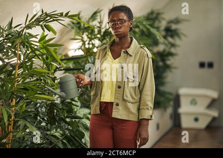 Portrait à la taille de la femme afro-américaine contemporaine qui arrose des plantes au bureau et regarde l'appareil photo, l'espace de copie Banque D'Images