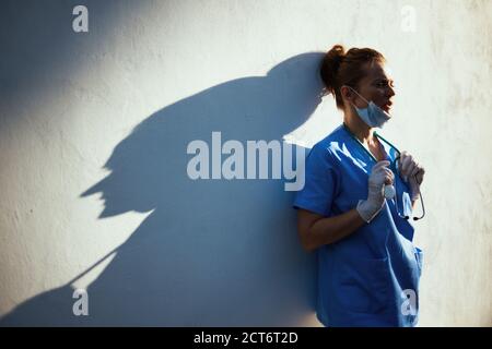 pandémie covid-19. femme médecin moderne fatiguée en exfoliant avec stéthoscope, masque médical et gants en caoutchouc à l'extérieur près du mur. Banque D'Images