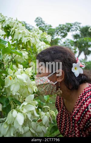 Belle dame avec masque floral, boucles d'oreilles et haut de créateur sent blanc fleurs de Mussaenda. Protection contre le corona avec élégance. Banque D'Images