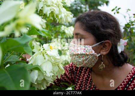 Belle dame avec masque floral, boucles d'oreilles et haut de créateur sent blanc fleurs de Mussaenda. Protection contre le corona avec élégance. Banque D'Images
