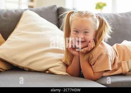 Portrait de petite fille avec le syndrome de Down sourire heureux à l'appareil photo tout en étant allongé sur le canapé à la maison, espace de copie Banque D'Images