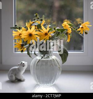 Un bouquet d'artichaut de jérusalem jaune dans un vase transparent sur un rebord de fenêtre blanc devant le jardin d'automne. Figurine en porcelaine d'un ours polaire. Banque D'Images