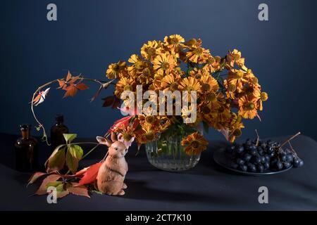 Un bouquet d'helenium orange avec des feuilles de raisin sauvage dans un vase en verre cannelé contre un mur bleu foncé. Figurine en porcelaine d'un lièvre. Banque D'Images