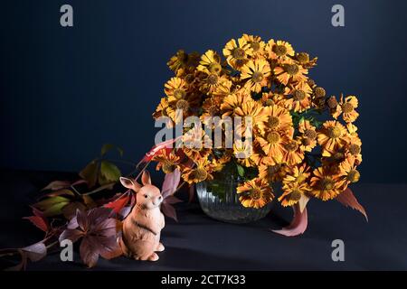Un bouquet d'helenium orange avec des feuilles de raisin sauvage dans un vase en verre cannelé contre un mur bleu foncé. Figurine en porcelaine d'un lièvre. Banque D'Images