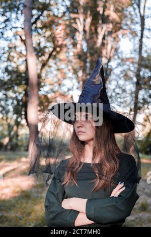 Jolie jeune belle femme en robe sombre et chapeau de sorcière debout au milieu des bois d'automne ou du parc. Costume de fête d'Halloween. Gros plan portrai Banque D'Images