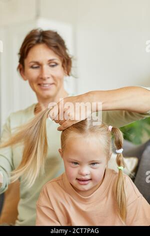 Portrait vertical de la mère souriante peignant les cheveux de la jolie fille avec le syndrome de down et de l'attacher dans les pigeons Banque D'Images