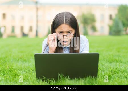 Oh non. Retour à l'école. Jeune fille utilise un ordinateur sur l'herbe verte dans le parc. Enfant dans des lunettes avec ordinateur portable. Nouvelle technologie dans la vie moderne. Étude. Enfant surpris travaillant sur ordinateur portable. Éducation en ligne. Banque D'Images
