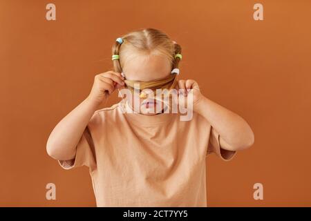 Portrait de petite fille avec le syndrome de Down cachant son visage tout en se tenant contre fond brun Uni dans le studio, espace de copie Banque D'Images