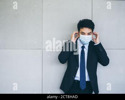 Jeune homme d'affaires asiatique en costume portant un masque médical sur fond de mur gris dans le bureau avec espace de copie, nouveau style de vie de travail normal après corona Banque D'Images