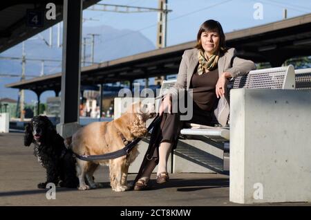 Femme élégante assise sur un banc dans une gare avec ses deux chiens. Banque D'Images