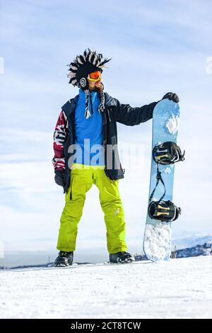 Bonhomme de neige à barbe dans un masque de ski avec des lunettes et un gros chapeau mohawk en fourrure sur fond de ciel et de montagnes enneigées d'hiver Banque D'Images