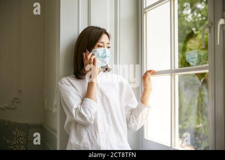 jeune femme asiatique en quarantaine portant un masque debout près de la fenêtre parler sur un téléphone portable a l'air triste et déprimé Banque D'Images