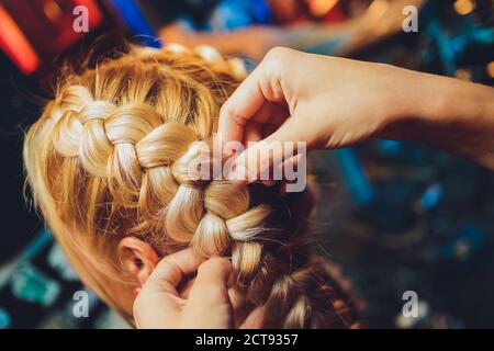 Processus de tressage le maître tisse des tresses sur sa tête blonde petite fille dans le salon de beauté gros plan. Soins professionnels des cheveux et création de coiffures. Banque D'Images