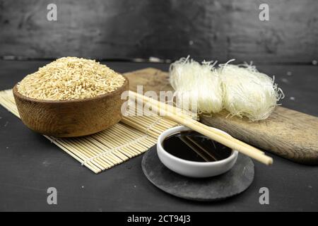 faites dorer le riz dans un bol en bois, cuisine asiatique avec tapis de bambou, baguettes, sauce soja, gingembre mariné et nouilles de riz en verre sur fond noir Banque D'Images