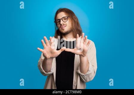 Homme caucasien long à poil long qui gestait un rejet avec des paumes portant des lunettes et la barbe sur un mur bleu de studio Banque D'Images