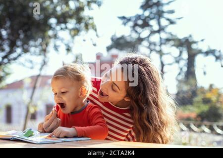 Happy Mother and Toddler Reading Lift-the-rabat Childrens Book and Eating En-cas dans le jardin Banque D'Images