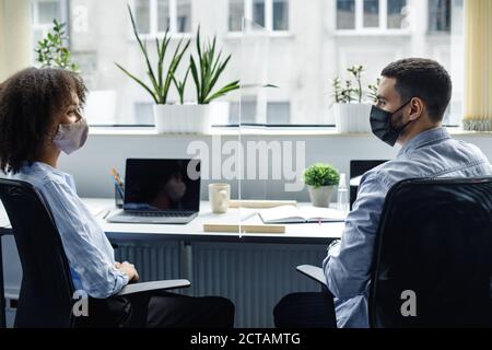 Femme afro-américaine et homme arabe dans les masques de protection communiquent, assis sur le lieu de travail avec des ordinateurs portables à l'intérieur Banque D'Images