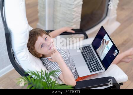 Un garçon heureux s'assoit sur une chaise avec un ordinateur portable pour passer un appel vidéo avec un chiot. Enfant parlant à son père et à son chien lors d'un chat vidéo. Banque D'Images