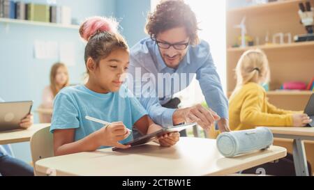 École élémentaire classe informatique : Cute Girl utilise un ordinateur numérique tablette, un professeur amical l'aide avec l'affectation. Enfants Getting Banque D'Images