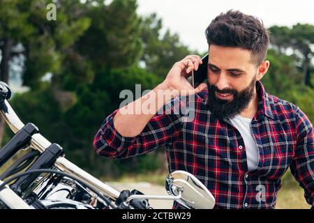 portrait d'un beau barbu parlant sur son téléphone portable assis sur sa moto, concept de liberté et de style de vie de motard, espace de copie pour le texte Banque D'Images