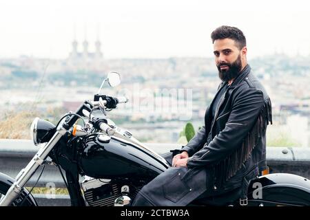 homme barbu habillé de cuir assis sur sa moto avec vue panoramique sur la ville, concept de liberté et de style de vie de motard, espace de copie pour le texte Banque D'Images