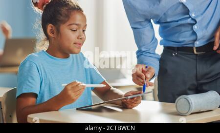 Cours de sciences informatiques de l'école élémentaire : Smart Girl utilise un ordinateur numérique tablette, un professeur amical l'aide en expliquant la leçon et l'envoi Banque D'Images