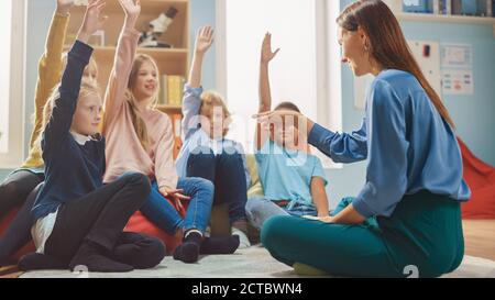 Cours de créativité à l'école élémentaire : les enfants assis sur le tapis tandis que l'enseignant explique la leçon et pose la question, chaque enfant soulève la main avec Banque D'Images