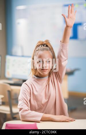 École primaire salle de classe: Portrait d'une petite fille mignonne avec des cheveux blond et queue de cheval levant la main avec une réponse. Un jeune étudiant brillant demande Banque D'Images
