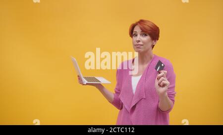 Une femme d'âge moyen à tête rouge confuse tenant un ordinateur portable et une carte de crédit. Concept d'achat en ligne. Isolé sur fond orange. Photo de haute qualité Banque D'Images