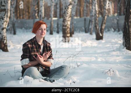 Ginger femme assise à la neige près de l'arbre. Jeune femme dans le parc d'hiver. Liberté et concept joyeux avec espace de copie. Banque D'Images