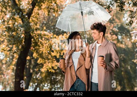 couple en trench-coats debout sous un parapluie et tenant le papier tasses avec café à emporter Banque D'Images